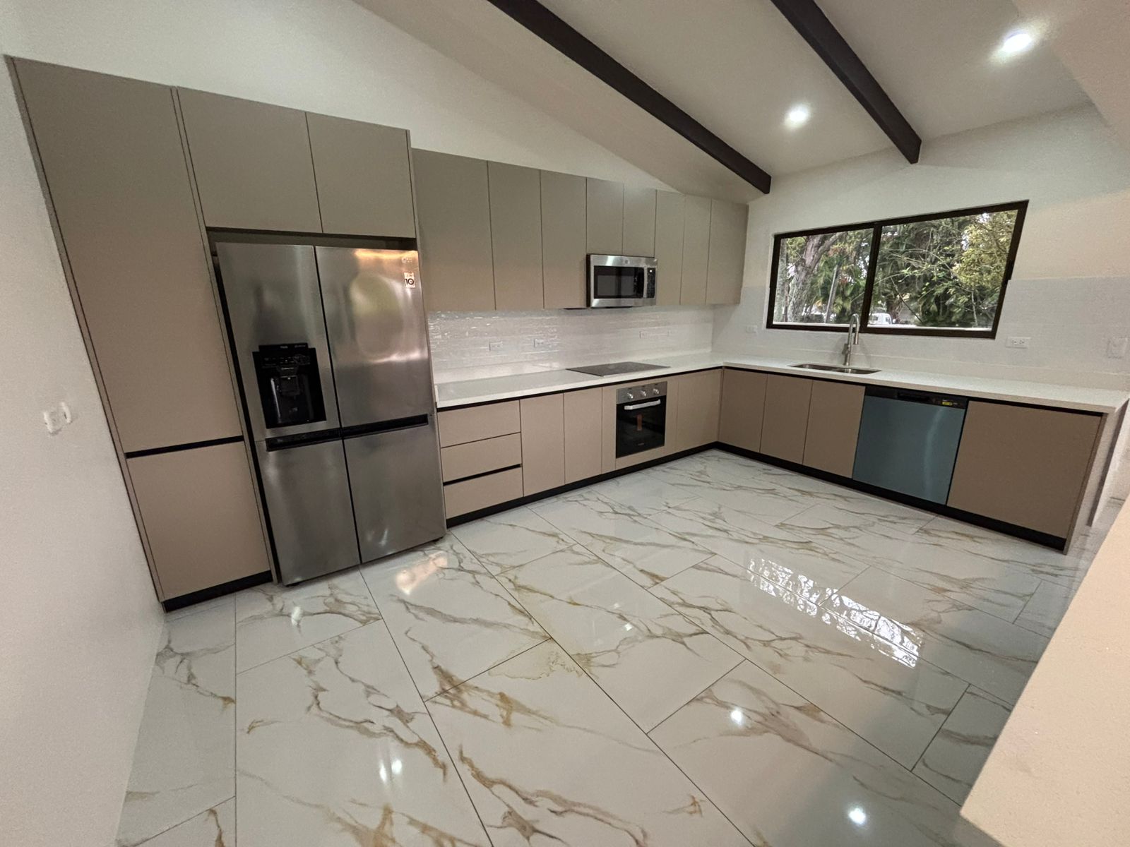 Cocina en L de tonos arena con piso marmoleado, vigas oscuras y gran ventanal lateral.
