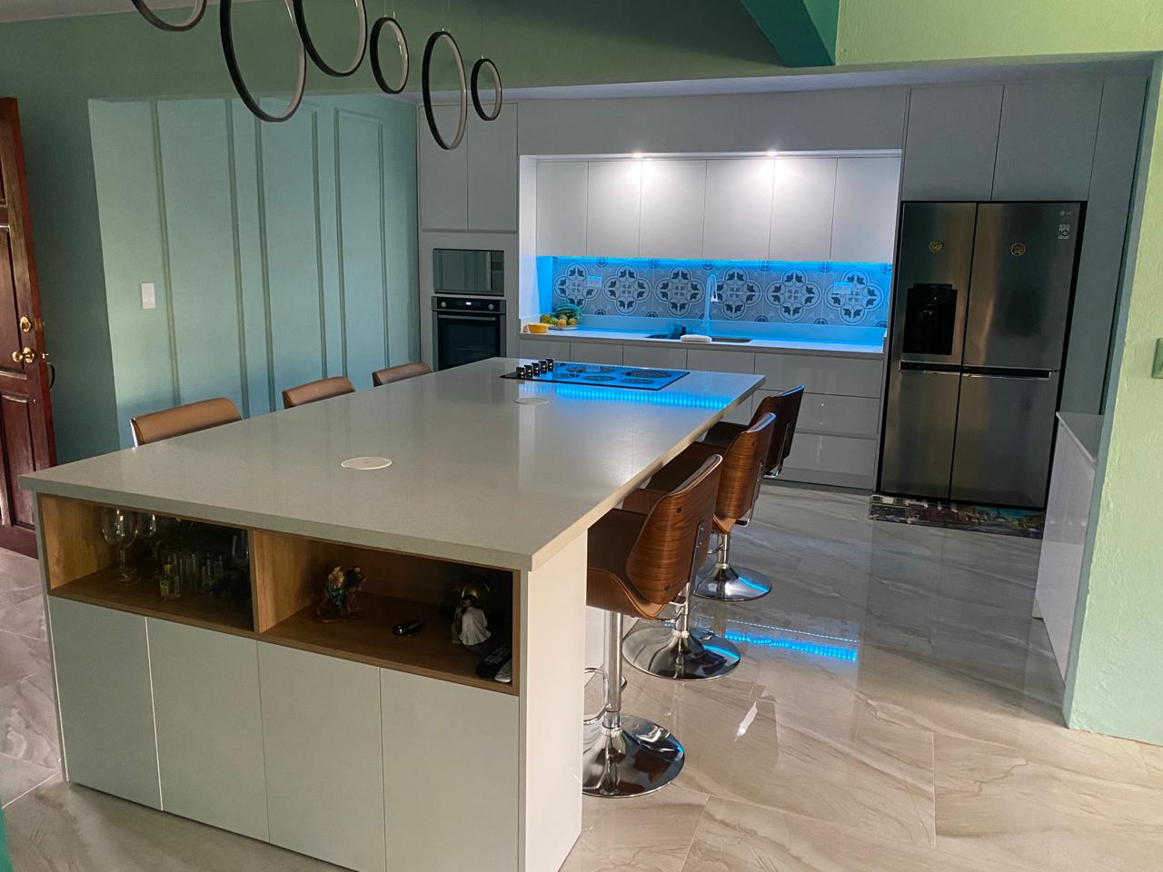 Cocina abierta con isla amplia, mobiliario claro y backsplash decorativo iluminado en azul.